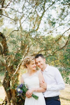 The groom gently hugs the bride near the green trees in the olive grove, the bride holds a bouquet of blue flowers . High quality photo