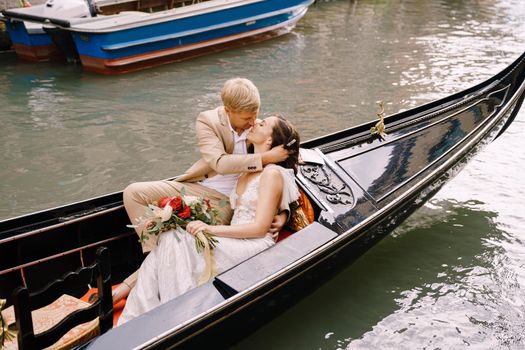 The bride and groom ride in a classic wooden gondola along a narrow Venetian canal. A close-up of cuddling newlyweds.