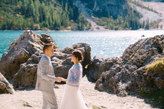 Bride and groom stand against the backdrop of stones overlooking Lago di Braies in Italy. Destination wedding in Europe, at Braies lake. In love newlyweds walk against the backdrop of amazing nature.