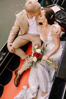 The gondolier rides the bride and groom in a classic wooden gondola along a narrow Venetian canal. Newlyweds sit in a boat against the background of ancient buildings.
