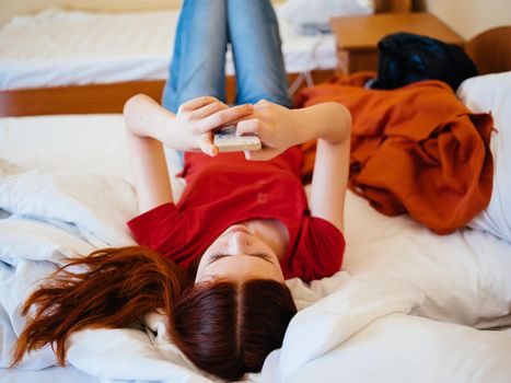 Happy woman in a red T-shirt and jeans lies in bed with a mobile phone in her hands. High quality photo