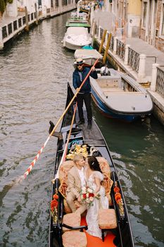 The gondolier rides the bride and groom in a classic wooden gondola along a narrow Venetian canal. Newlyweds sit in a boat against the background of ancient buildings.