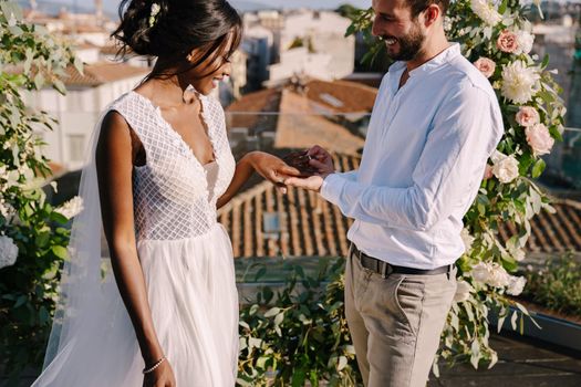 Interracial wedding couple - African-American bride and Caucasian groom. Groom puts a ring on brides finger. Destination fine-art wedding in Florence, Italy. Wedding ceremony on the roof of building