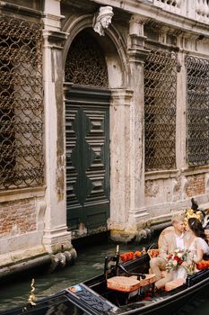 The gondolier rides the bride and groom in a classic wooden gondola along a narrow Venetian canal. Newlyweds sit in a boat spout to the spout, swim against the background of an old forged lattice.