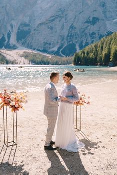 The groom dons a bride's ring, in place for the ceremony, with an arch of autumn floral columns, against the backdrop of Lago di Braies in Italy. Destination wedding in Europe, at Braies lake.