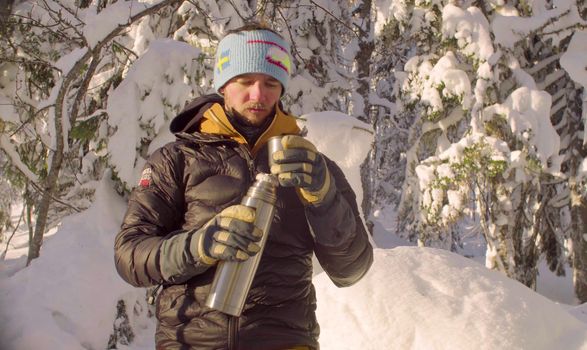 Young man drinking hot tea from a thermos in winter forest