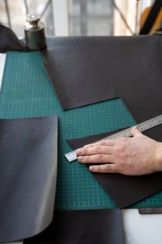 Men's hand holding a stationery knife and metal ruler and cutting on a pieces for a leather wallet in his workshop. Working process with a brown natural leather. Craftsman holding a crafting tools