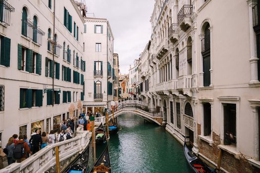 A crowd of tourists walks the streets of Venice, in Italy, near narrow canal between buildings. Small arched bridges over sea water