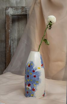 White flower in Blue and White, Red, Yellow handmade ceramic vase on Blush textured table cloth. Home decor, Copy space, Selective focus.