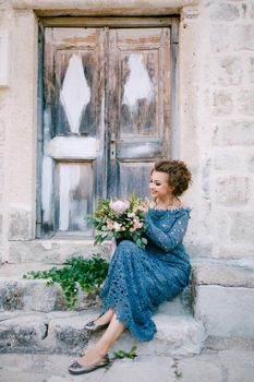 A bride in a stylish blue dress with a bouquet in her hands sits on the steps near an old wooden door . High quality photo