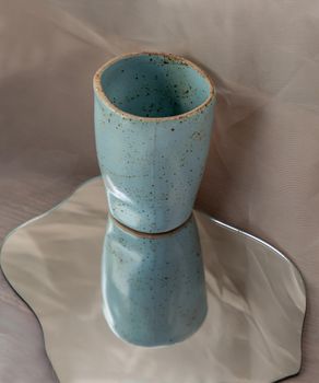 Blue handmade ceramic mug on mirror on fabric textured table cloth. Home decor, Copy space, Selective focus.