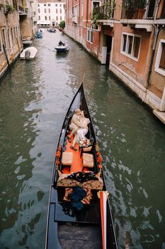 The gondolier rides the bride and groom in a classic wooden gondola along a narrow Venetian canal. Newlyweds sit in a boat against the background of ancient buildings.