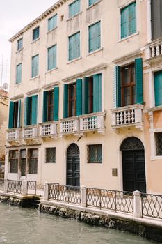 The four-storey building is short-colored with green forged wooden shutters and balconies with white columns. A beautiful metal forged fence near the water.