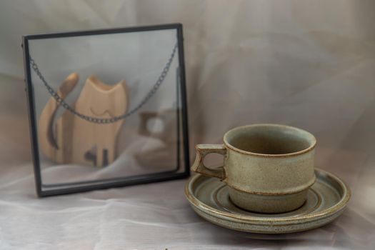 Handmade ceramic coffee mug with wooden doll in cat shape on fabric textured table cloth. Home decor, Copy space, Selective focus.