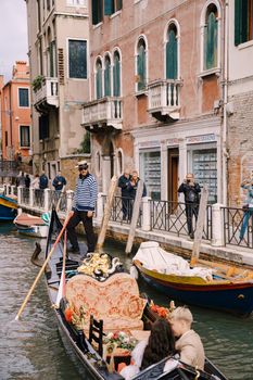 The gondolier rides the bride and groom in a classic wooden gondola along a narrow Venetian canal. Newlyweds sit in a boat and kiss.