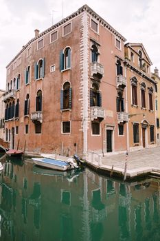 A wide frame of the corner of the three-story house, peach-colored, near a small narrow Venetian Canal. A motorboat moored near the walls of a house standing in the water in Venice, Italy.