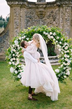 The groom is circling the bride in her arms. Round wedding arch decorated with white flowers and greenery in front of an ancient Italian architecture. Wedding at an old winery villa in Tuscany, Italy.