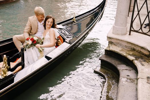 The bride and groom ride in a classic wooden gondola along a narrow Venetian canal. A close-up of cuddling newlyweds.