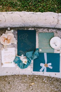 Blue mockup of invitations to wedding, blue envelope with seal print of sealing wax, a silk blue ribbon, a white sheet with the colors of dess code, pink flower against background of marble stone