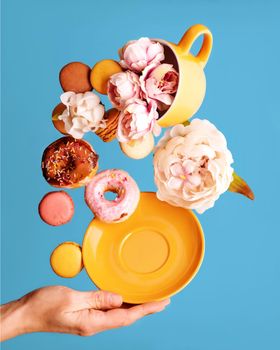 still life of peons in yellow cup, donuts, macaroons, peony and hand holding saucer flying over a blue background