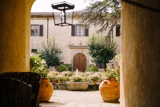 Fountain surrounded by flowerpots in front of the old villa in Italy, in Tuscany, near Florence. Facade of the building with an open door under the shade of trees.