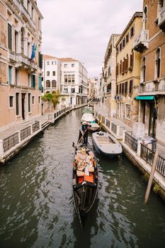 The gondolier rides the bride and groom in a classic wooden gondola along a narrow Venetian canal. Newlyweds sit in a boat against the background of ancient buildings.