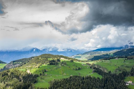Aerial view of improbable green meadows of the Italian Alps, green slopes of the mountains, Bolzano, huge clouds over a valley, roof tops of houses, Dolomites on background, sunshines through clouds