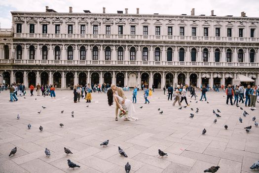 Wedding in Venice, Italy. The bride and groom kiss among the many pigeons in Piazza San Marco, against backdrop of the National Archaeological Museum Venice, surrounded by a crowd of tourists.