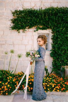 A bride in a blue dress with a bouquet stands at the wall of a house with a green liana and orange flowers, close-up. High quality photo