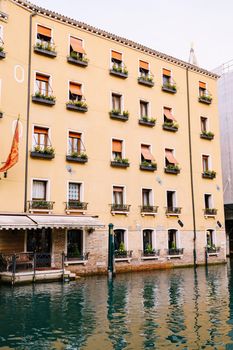 Yellow five-story building in Venice, Italy. The main entrance of the building goes into the water, at entrance there is a sofa, on horseback there are flower pots and orange shutters from the sun
