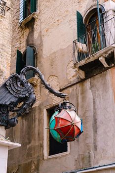 An unusual street lamp in the shape of The Maforio Dragon on the corner of the building, with a ball of three umbrellas - blue, green and red, on the streets of Venice, Italy, near Rialto bridge.