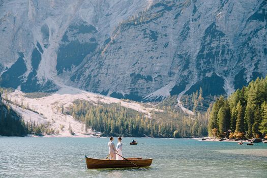 The bride and groom swim in a wooden boat with oars at Lago di Braies in Italy. Wedding in Europe, at Braies lake. Newlyweds standing hugging in the boat.