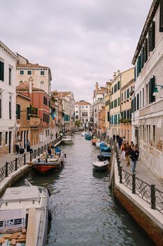 A classic view of the narrow sea Venetian canal. Colorful buildings stand opposite each other, boats moored near the shore, gondolier rides tourists on a gondola. Blue skies on a sunny day.