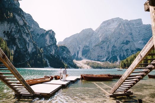 The bride and groom walk on a wooden boat dock at Lago di Braies in Italy. Wedding in Europe, at Braies lake. Newlyweds walk, kiss, cuddle against the backdrop of rocky mountains.