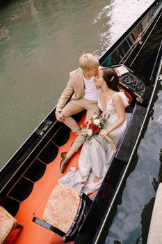 The gondolier rides the bride and groom in a classic wooden gondola along a narrow Venetian canal. Newlyweds sit in a boat against the background of ancient buildings.