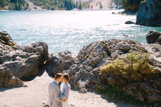 Bride and groom stand against the backdrop of stones overlooking Lago di Braies in Italy. Destination wedding in Europe, at Braies lake. In love newlyweds walk against the backdrop of amazing nature.