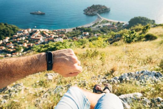 Fitness bracelet on a man's hand. A man sits on top of a mountain overlooking the island of Sveti Stefan in Montenegro