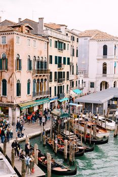 Gondola pier near the Rialto Bridge, Venice, Italy, Europe. Tourists walk on souvenir shops, stand in line for tour on a gondola. A lot of people on promenade, against background of ancient building