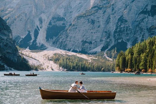 Bride and groom swim in wooden boat, on Lago di Braies lake in Italy. Wedding in Europe - Newlyweds sitting in boat and kissing.