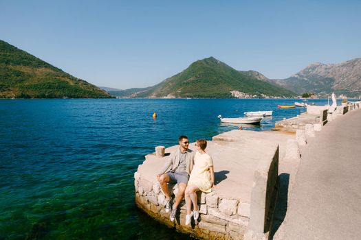 A man and a woman sit side by side on the edge of a pier near Perast in the Bay of Kotor . High quality photo