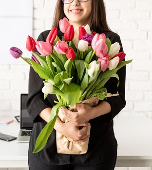 Small business concept. Portrait of young confident brunette business woman standing by the desk with fresh tulips bouquet