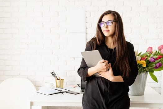 Small business concept. Portrait of young confident brunette business woman standing by the desk with fresh tulips holding digital tablet