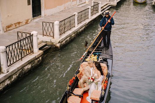 The gondolier rides the bride and groom in a classic wooden gondola along a narrow Venetian canal. Newlyweds sit in a boat against the background of ancient buildings.