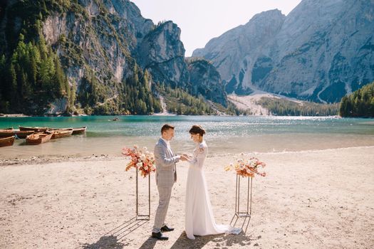 The bride dons a ring to the groom at the venue for the ceremony, with an arch of autumn floral columns, against the backdrop of Lago di Braies in Italy. Destination wedding in Europe, at Braies lake.
