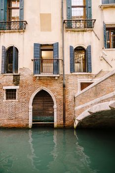 A brick building in the water in Italy, Venice. A brick bridge over a small narrow canal, classic Venetian windows in the facade of building with wooden shutters and forged bars.