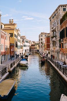 A classic view of the narrow sea Venetian canal. Colorful buildings stand opposite each other, boats moored near the shore, gondolier rides tourists on a gondola. Blue skies on a sunny day.