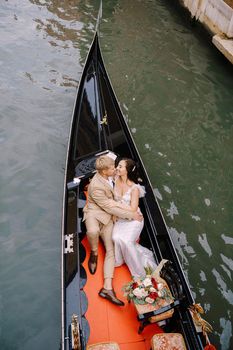 The gondolier rides the bride and groom in a classic wooden gondola along a narrow Venetian canal. Newlyweds sit in a boat against the background of ancient buildings.