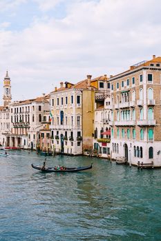 A gondola with tourists is sailing along Grand Canal amid the facades of Venetian houses standing on the water in Venice, Italy.
