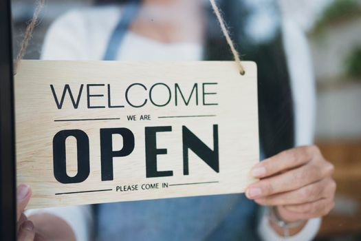 Small business owner smiling while turning the sign for the reopening of the place after the quarantine due to covid-19. Close up of woman  hands holding sign now we are open support local business.