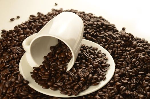 An abundant pile of coffee beans surrounds a white cup and saucer over a clean white background.